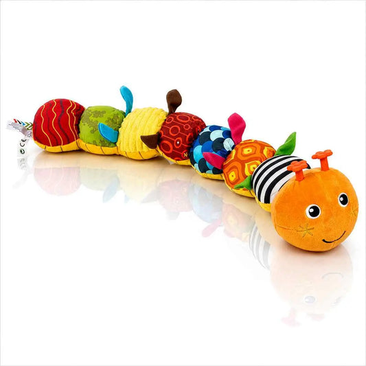 "A long, colorful plush caterpillar toy rests on a reflective white surface. The toy features an orange head with a smiling face and orange antennae, followed by several rounded body segments in various vibrant colors and patterns, including red swirls, green floral, yellow corduroy, and black-and-white stripes. Small, soft fabric fins or legs in blue, brown, pink, and green protrude from the sides of the segments."
