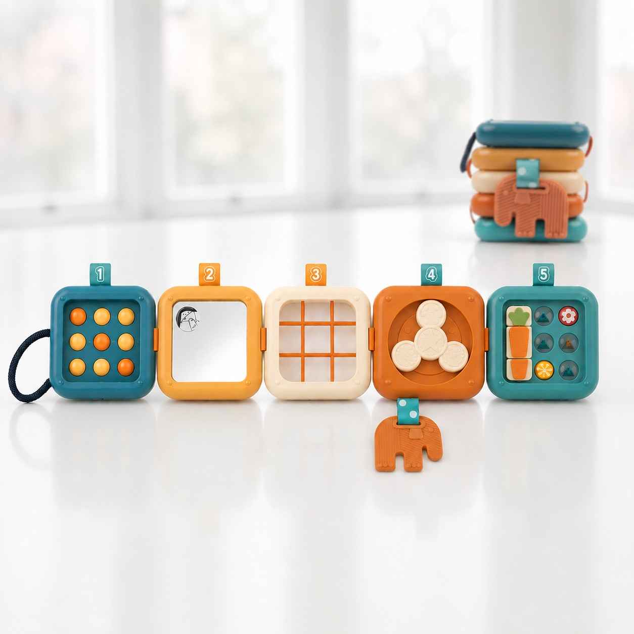 A colorful five-piece toddler stacking toy displayed on a white surface. Five square, interlocking blocks numbered 1 to 5 are laid out horizontally in a row, each featuring a different sensory activity like buttons, a mirror, and a spinner. A small orange elephant tag hangs from block number 4, while a stacked version of the identical toy sits blurred in the background.
